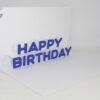 A greeting card with a white base, with a white insert that has the words Happy Birthday that pop up. Individual letters spelling Happy Birthday in blue are glued to the white insert of the card. Card is shown at an angle, so the pop up can be seen
