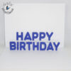 A greeting card with a white base, with a white insert that has the words Happy Birthday that pop up. Individual letters spelling Happy Birthday in blue are glued to the white insert of the card.